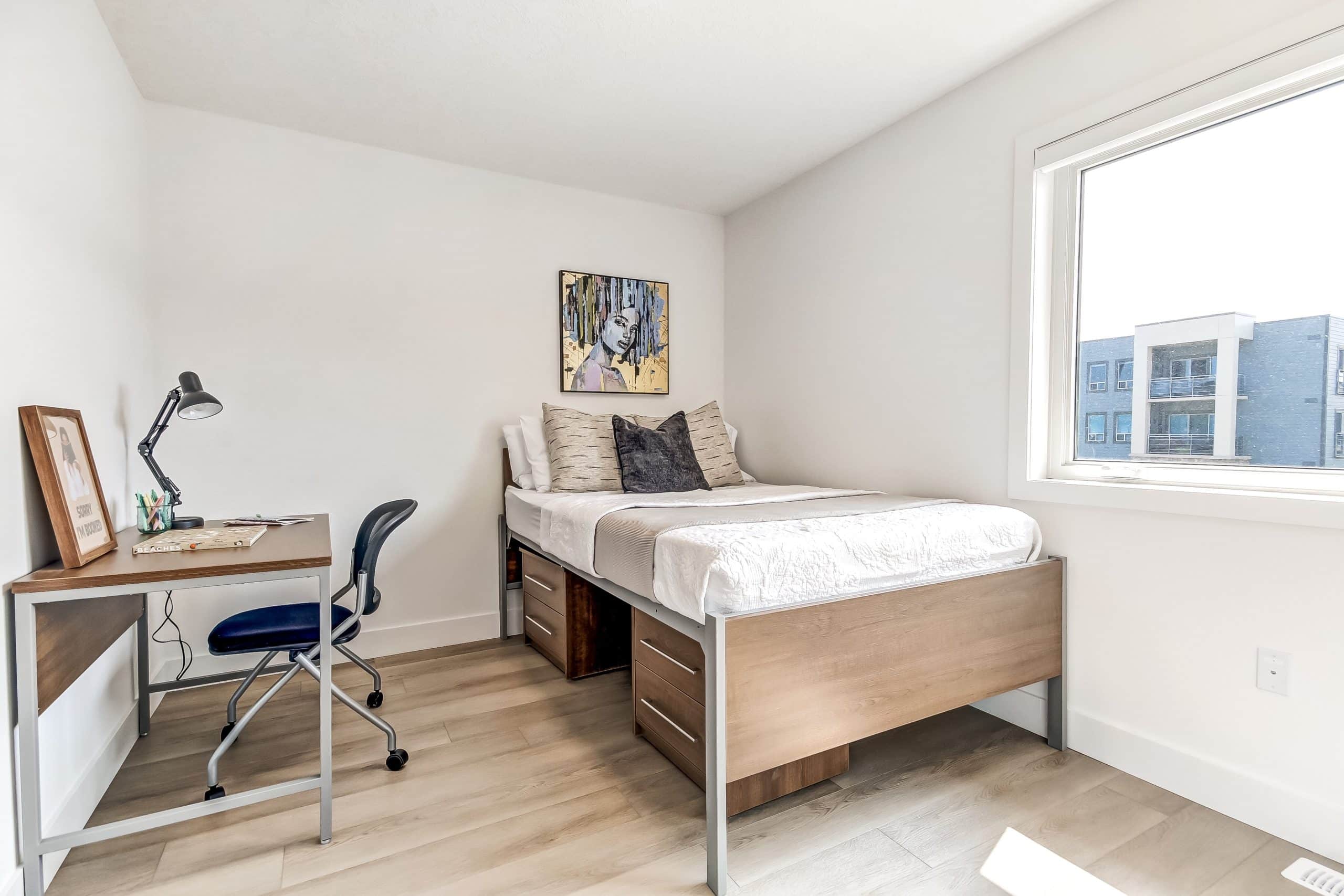 Bright minimalist bedroom with a neatly made bed, desk and blue chair, abstract wall art, and a large window overlooking modern buildings.